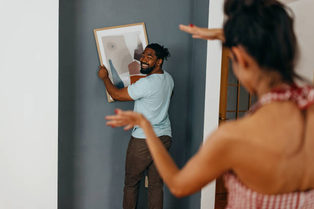 Level it to the right Black man holding framed picture while Caucasian woman is helping to level it against a the wall decorating stock pictures, royalty-free photos & images