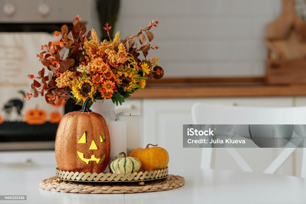 A vase of flowers,a jack pumpkin and candles on a tray. In the background - the interior of a white kitchen in Scandi style. The concept of home and comfort. Autumn decor for the Halloween holiday. A postcard, a flyer for a party, a Halloween holiday. Bright pumpkins on a light background. Autumn Stock Photo