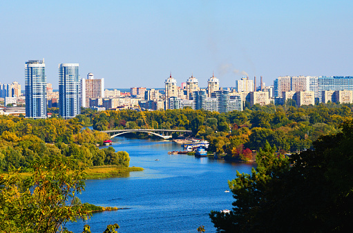Scenic autumn landscape view of Kyiv. Nature landscape with autumn colors. Dnipro River and Hydropark against blue sky. Modern colorful residential skyscrapers in the background. Kyiv, Ukraine.