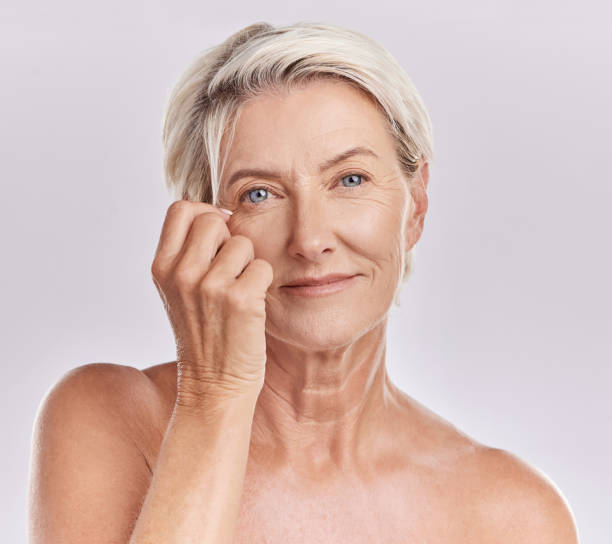 portrait d’une femme caucasienne mature et souriante et heureuse, à l’air positive et joyeuse tout en prenant soin de sa peau dans un studio sur fond de copyspace violet. femme âgée faisant sa routine de soins de la peau - real people indoors studio shot head and shoulders photos et images de collection