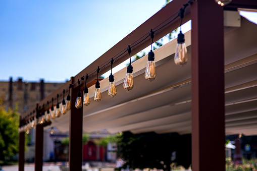 textile awning in city street cafe wooden frame gazebo with garland of strings of retro edison light bulbs glow with warm light on touristic summer day close-up cityscape, nobody.