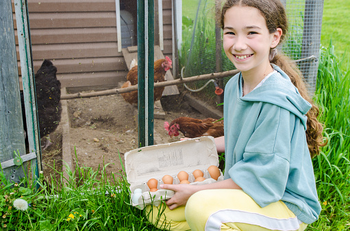 Girl holding fresh eggs in front of chicken coop in backyard of farmland