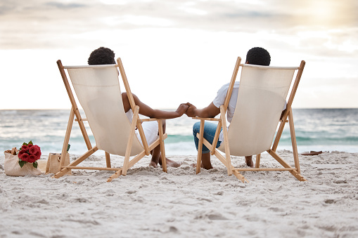 Rearview of an african American couple spending a day at the sea together. Content boyfriend and girlfriend holding hands while sitting on the beach. Caring husband and wife bonding on the seashore