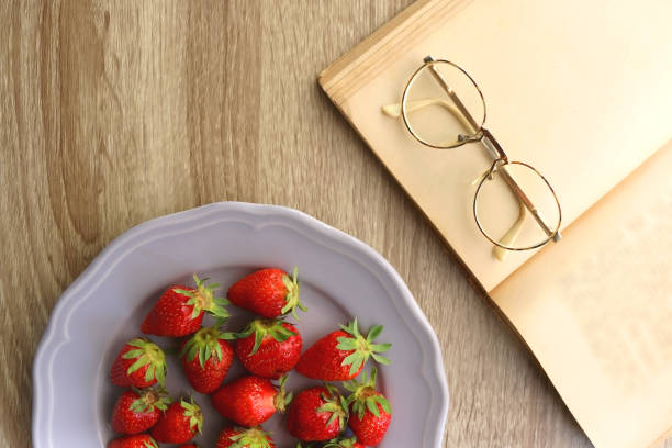 Lilac plate full of fresh strawberries, open book and reading glasses on wooden table. Flat lay.