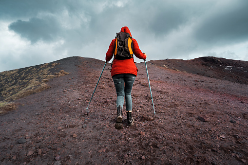 A young woman hiker climbs a rocky terrain of lava towards the volcano Mount Etna in Sicily, Italy, with a backpack behind her, helping herself with trekking poles in cloudy day