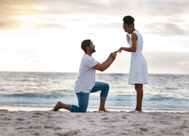 novio pidiéndole a su novia que se case con él mientras están juntos en la playa. hombre afroamericano proponiéndole matrimonio a su novia a la orilla del mar. joven pareja feliz que se compromete de vacaciones - prometido fotografías e imágenes de stock