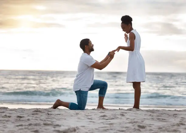 Photo of Boyfriend asking his girlfriend to marry him while standing on the beach together. African american man proposing to his girlfriend by the seashore. Young happy couple getting engaged on holiday