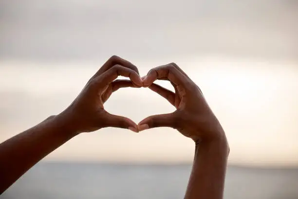 Closeup of the hands on an african american female making a heart shape against the sky outdoors. Confident black woman standing on a beach outside at sunset gesturing toward the sea. Health and love