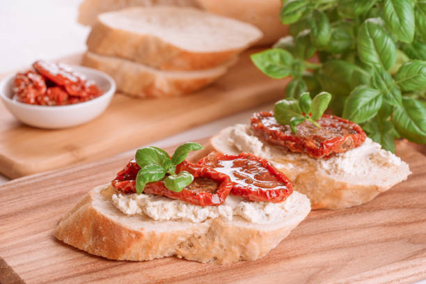 concepto de desayuno casero saludable. deliciosas tostadas con queso ricota, tomates secados al sol con aceite de oliva rematados con albahaca sobre tabla de cortar de madera. aperitivo clásico italiano - brushetta fotografías e imágenes de stock