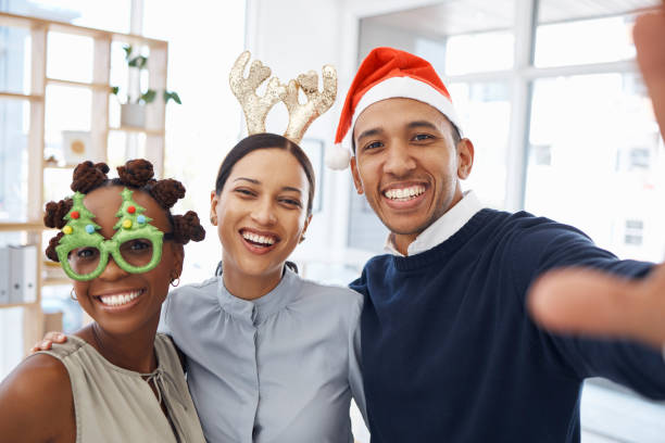 Group of three cheerful diverse businesspeople taking a selfie together at work. Happy mixed race businessman taking a photo with his joyful female colleagues at a Christmas office party Group of three cheerful diverse businesspeople taking a selfie together at work. Happy mixed race businessman taking a photo with his joyful female colleagues at a Christmas office party office christmas party stock pictures, royalty-free photos & images