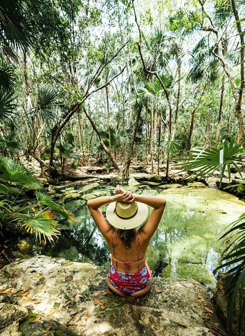 young hispanic woman with hat enjoys looking at a cenote in mexico