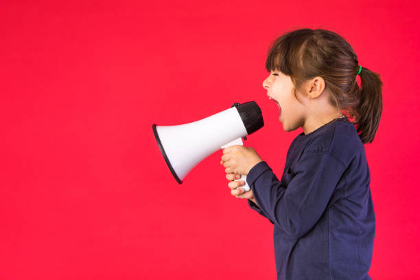 Black-haired girl in a blue sweater and white pants, shouting through a white megaphone, on a red background. stock photo