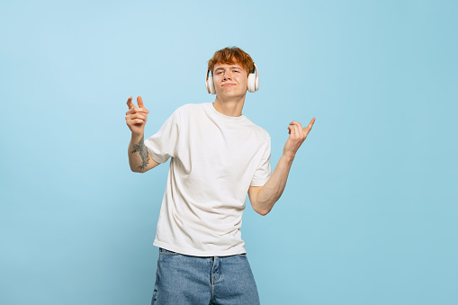 Listening to music. Studio shot of young red-headed man in white t-shirt posing isolated over blue studio background. Concept of youth, fashion, emotions, facial expression. Copy space for ad