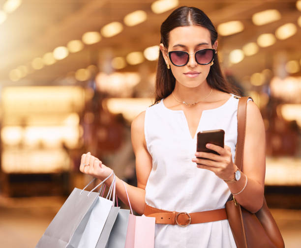 giovane donna alla moda che indossa occhiali da sole usando un telefono cellulare mentre trasporta borse durante uno shopping. giovane donna bruna che invia un messaggio con il suo smartphone dopo aver fatto shopping in un centro commerciale. donna che con - shopping mall retail shopping sale foto e immagini stock