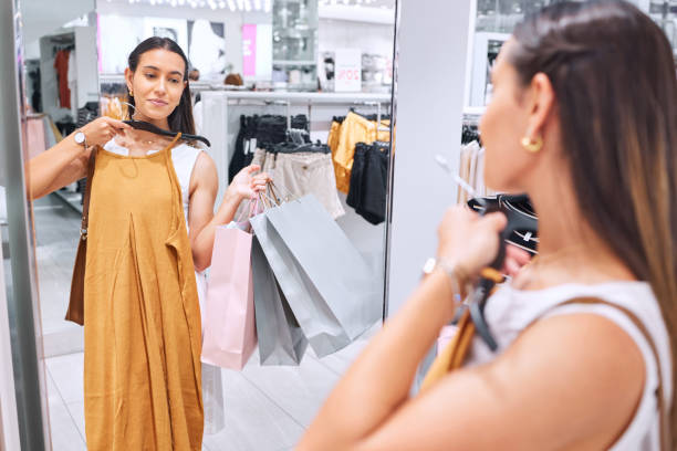 백화점의 거울에 담긴 옷을 보고 있는 아름다운 젊은 여성. 패션 판매를 위해 쇼핑하는 동안 옷을 입으려고합니다. 소매 치료는 그녀가 필요로하는 것입니다. 고객은 항상 옳습니다. - mirror women dress looking 뉴스 사진 이미지