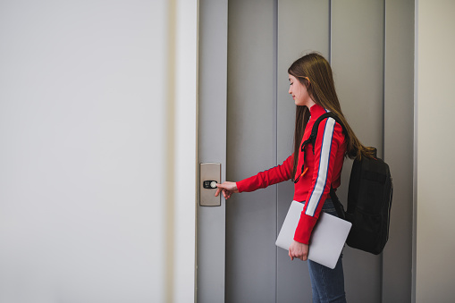 Young girl with backpack and laptop standing near elevator door, pressing call button.