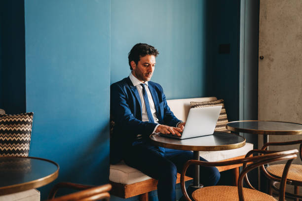 あるビジネスマンがカフェのテーブルに座りながらノートパソコンを使って作業している - business blue business person businessman ストックフォトと画像