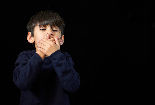 serious brunette little boy looking at the camera covering his mouth with both hands. black background and copy space. Concept of silence or secret. horizontal