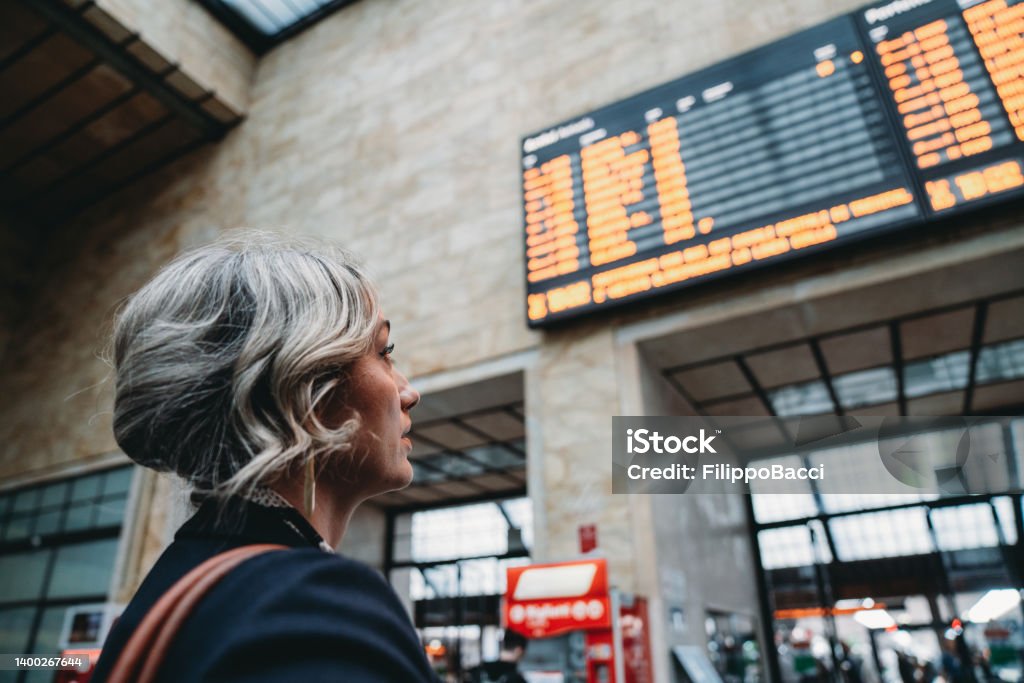 Rear view of a woman checking the arrival departure board in a railroad station Rear view of a woman checking the arrival departure board in a railroad station. Selective focus on the woman. Railroad Station Stock Photo