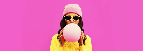 Fashionable portrait of stylish cool young woman inflating chewing gum wearing yellow knitted sweater, hat on pink background, blank copy space for advertising text stock photo