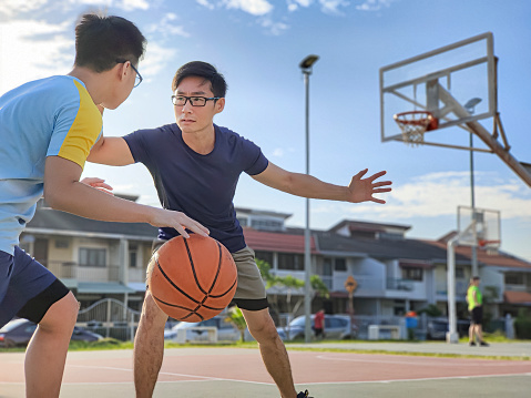Asian chinese father and son having fun, playing basketball outdoors during weekend morning