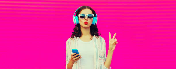 Portrait of young woman in headphones listening to music with smartphone on pink background, blank copy space for advertising text stock photo