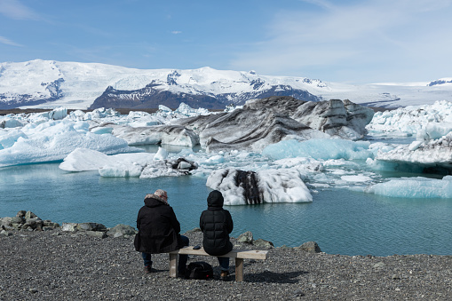 Two people sitting at Jokulsarlon, a glacial lagoon with icebergs in southern Iceland. Jokulsarlon formed around 1935 and the size of the lake keeps increasing due to the melting glaciers.