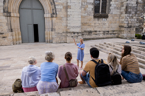 An over the shoulder view of a group of students sat on some stone steps and getting to know all about the historic town of village of Auvillar. Their teacher is stood in front of them and is taking questions from the group.