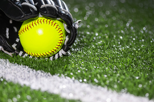 Selective focus on a new yellow softball in a black and white leather glove sitting on the turf next to the white baseline on an artificial turf during a night game.