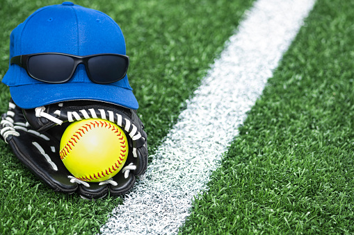 A blue baseball hat with dark sunglasses sitting atop a black and white glove with a new yellow softball in it, sitting along the white baseline during a day game