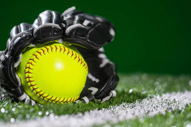 Selective focus on a new yellow softball in a black and white leather glove sitting on the turf next to the white baseline on an artificial turf during a night game.