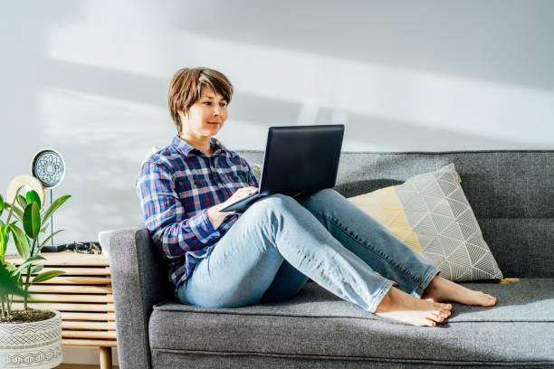 istock Smiled mixed race woman using laptop at home sitting on soft couch. Cozy workspace in modern interior with green plants. Remote work at home. Online education or entertainment. Copy space 1400277051