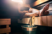 Sauna, steam room bath. Woman relaxing in spa. Wellness and warm temperature therapy in dark wood home in Finland. Water bucket and ladle. Towel on body, resting legs.