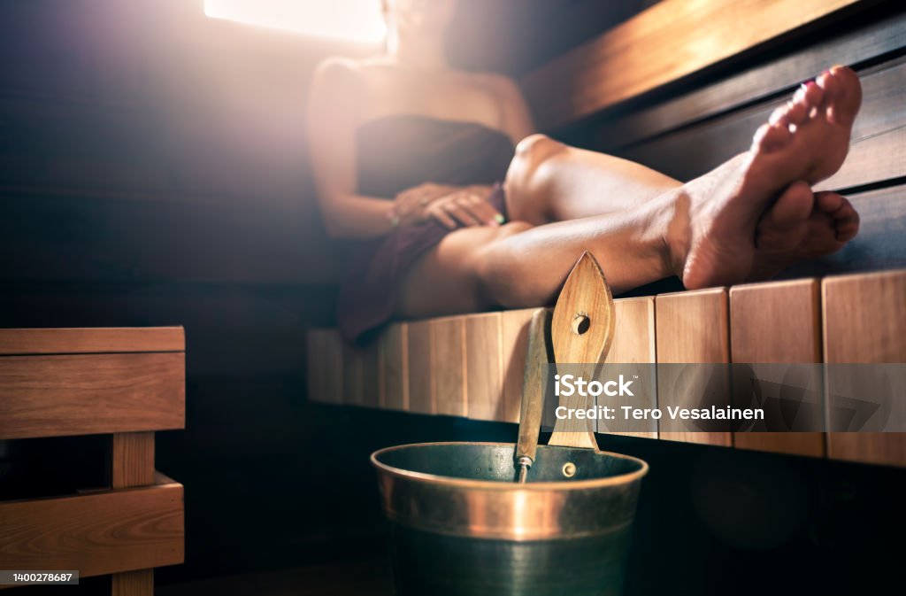 Sauna, steam room bath. Woman relaxing in spa. Wellness and warm temperature therapy in dark wood home in Finland. Water bucket and ladle. Towel on body, resting legs. Sauna, steam room bath. Woman relaxing in spa. Wellness and warm temperature therapy in dark wood home in Finland. Water bucket and ladle. Towel on body, resting legs. Traditional Finnish lifestyle. Sauna Stock Photo