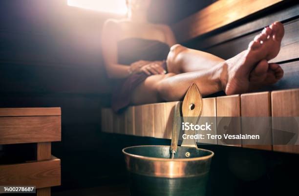 Sauna Steam Room Bath Woman Relaxing In Spa Wellness And Warm Temperature Therapy In Dark Wood Home In Finland Water Bucket And Ladle Towel On Body Resting Legs Stock Photo - Download Image Now