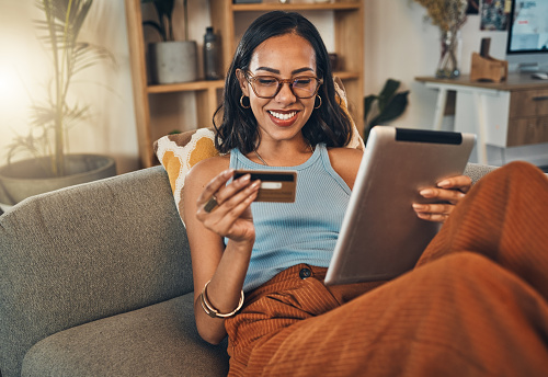 Mujer sonriente de raza mixta que usa tarjeta de crédito para el comercio electrónico en una tableta digital en casa. Feliz hispano sentado solo en el sofá de la sala de estar, utilizando la tecnología para la banca electrónica. Relajarse, ordenar, co photo