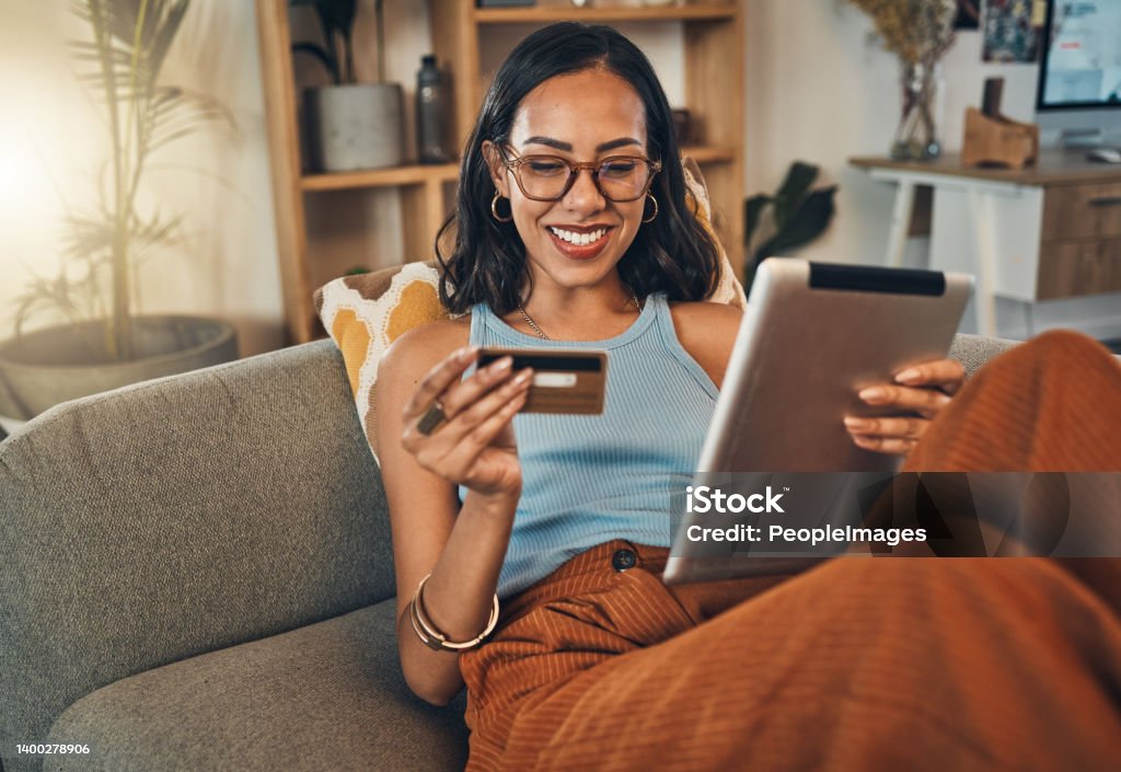 Smiling mixed race woman using credit card for ecommerce on digital tablet at home. Happy hispanic sitting alone on living room sofa, using technology for ebanking. Relaxing, ordering, buying online Credit Card Stock Photo