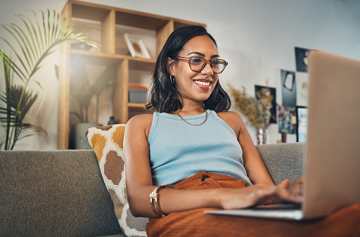 Mujer sonriente de raza mixta que navega por Internet en una computadora portátil en casa. Una feliz empresaria hispana sentada sola en su sofá y blogueando durante un fin de semana. Relajarse en una sala de estar y escribir una publicación de blog photo