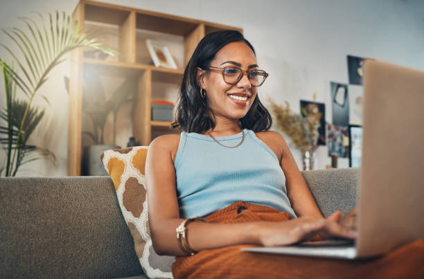 smiling mixed race woman browsing internet on a laptop at home. one happy hispanic entrepreneur sitting alone on her sofa and blogging over a weekend. relaxing in a living room and typing a blog post - working at home imagens e fotografias de stock