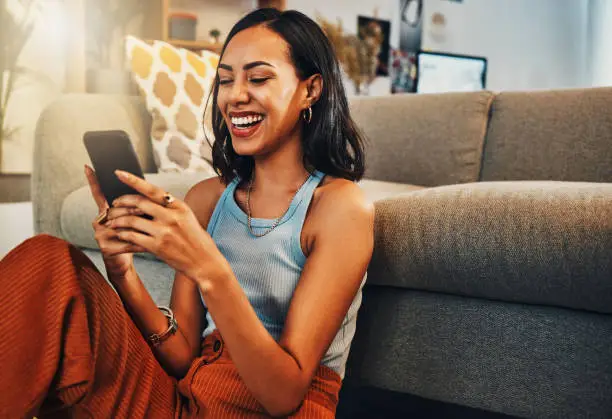 Photo of Beautiful mixed race woman browsing internet on cellphone in home living room. Happy hispanic sitting alone on floor in lounge and using technology to network. Laughing while scrolling on social media