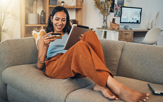 Full length smiling mixed race woman using credit card for ecommerce on digital tablet at home. Happy hispanic sitting alone on sofa, using technology for ebanking. Relaxing, ordering, buying online