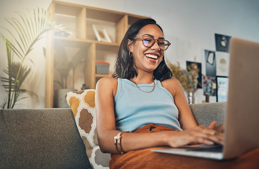 Smiling mixed race woman browsing internet on a laptop at home. One happy hispanic entrepreneur sitting alone on her sofa and blogging over a weekend. Relaxing in a living room and typing a blog post