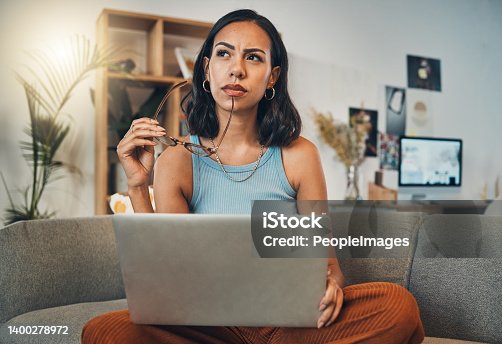 istock Beautiful mixed race woman thinking while using laptop for blogging in living room at home. Hispanic entrepreneur sitting cross legged alone on lounge sofa and planning next blog post on technology 1400278972