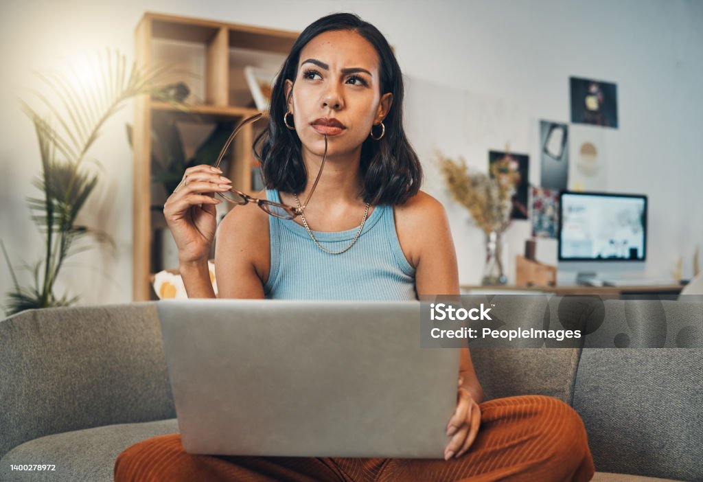 Beautiful mixed race woman thinking while using laptop for blogging in living room at home. Hispanic entrepreneur sitting cross legged alone on lounge sofa and planning next blog post on technology Contemplation Stock Photo