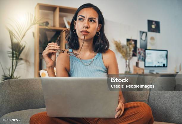 Beautiful Mixed Race Woman Thinking While Using Laptop For Blogging In Living Room At Home Hispanic Entrepreneur Sitting Cross Legged Alone On Lounge Sofa And Planning Next Blog Post On Technology Stock Photo - Download Image Now