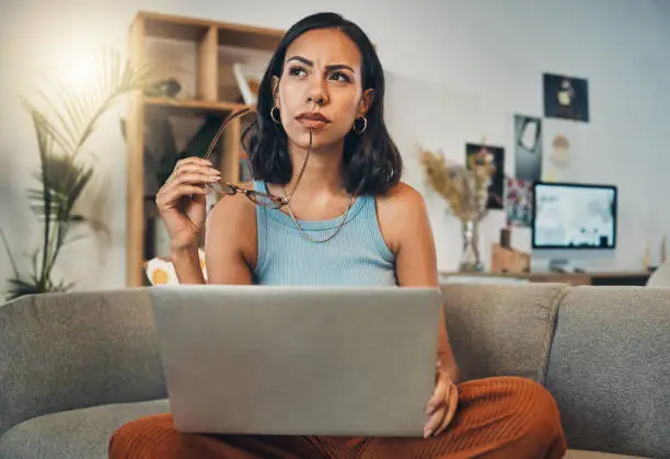 Beautiful mixed race woman thinking while using laptop for blogging in living room at home. Hispanic entrepreneur sitting cross legged alone on lounge sofa and planning next blog post on technology