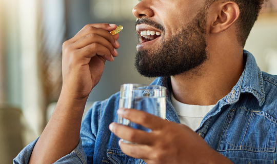 Bachelor drinking medication with a glass of water. Hispanic man drinking a supplement capsule in his apartment. Man using a glass of water to take his drugs. Sick man in recovery with a pill