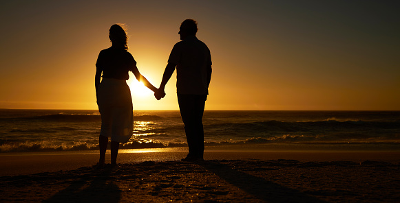 Silhouette of mature couple holding hands on the beach at sunset. Senior couple bonding on holiday together. Mature couple being affectionate on vacation on the beach. Older couple on seaside vacation