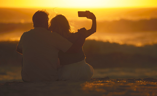 Back of a senior couple taking selfies on the beach. mature woman using a cellphone to take photos on the beach. Carefree mature couple taking photos with a smartphone. Couple enjoying a holiday