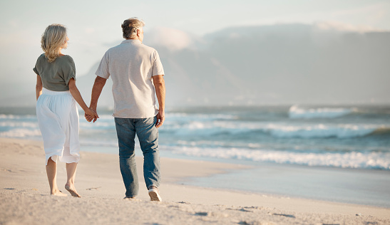 Mature couple walking on the beach. Back of senior couple holding hands on holiday together. Mature couple enjoying a holiday by the sea. Senior couple being affectionate and bonding on the beach